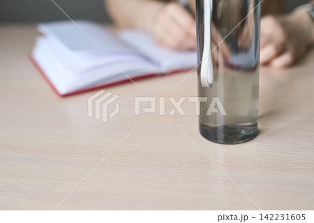 Glass Bottle Partially Filled On Wooden Table With Blurred Notebook And Person Writing Glass Bottle Partially Filled On Wooden Table With Blurred Notebook And Person Writing 142231605
