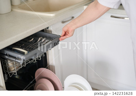 Person Sliding Dishwasher Cutlery Tray Outward Next To Sink And Countertop In White Kitchen While Person Sliding Dishwasher Cutlery Tray Outward Next To Sink And Countertop In White Kitchen While 142231628
