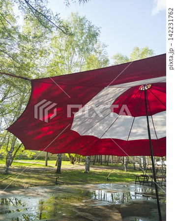 Large Red And White Patio Umbrella Open Above Wet Paved Area In Park With Reflected Puddles Large Red And White Patio Umbrella Open Above Wet Paved Area In Park With Reflected Puddles 142231762