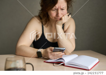 Woman Frowning While Reading Smartphone And Pausing With Pen Above Open Notebook At Wooden Table Woman Frowning While Reading Smartphone And Pausing With Pen Above Open Notebook At Wooden Table 142231777