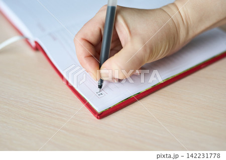 Child Hand Marking Checkbox In Red Bound Planner On Wooden Desk Close-Up, Pen Held Between Fingers Child Hand Marking Checkbox In Red Bound Planner On Wooden Desk Close-Up, Pen Held Between Fingers 142231778