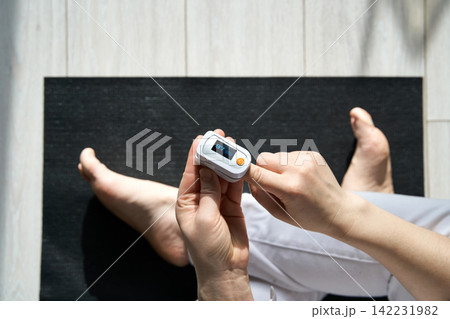 Person Checking Oxygen Level Using Pulse Oximeter While Sitting Barefoot On Yoga Mat On Light Wood 142231982