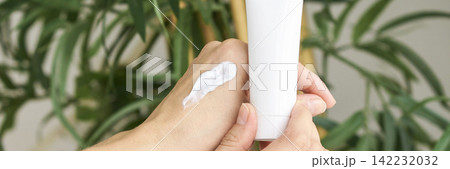 Close Up Of Hands Holding Cosmetic Tube With Cream On Skin Against Green Plant Background 142232032