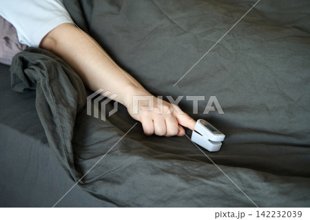 Person Reaching With Finger Toward Pulse Oximeter Placed On Bed Sheet While Partially Covered 142232039