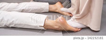 Practitioner Holding And Stretching Bare Foot Of Reclining Client On Mat Beside Seated Person Practitioner Holding And Stretching Bare Foot Of Reclining Client On Mat Beside Seated Person 142232181
