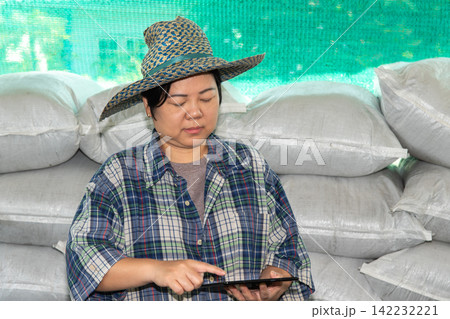 Female farmer wearing straw hat uses tablet beside stacked agricultural sacks representing smart farming digital technology crop management modern agriculture 142232221