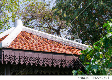 Traditional Thai roof terracotta tiles decorative wooden eaves surrounded by lush green trees showing historic architecture cultural heritage tropical craftsmanship 142232391