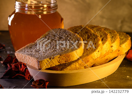 Golden honey and fresh bread slices beside glass jar in warm rustic kitchen setting Golden honey and fresh bread slices beside glass jar in warm rustic kitchen setting 142232779