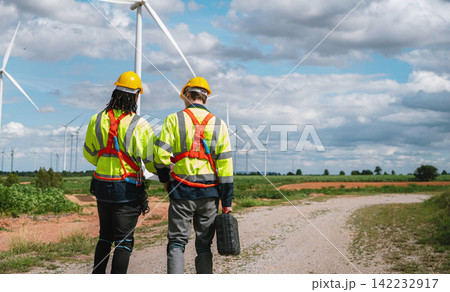 Two engineers walk toward wind turbines under a partly cloudy sky Two engineers walk toward wind turbines under a partly cloudy sky 142232917