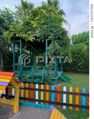 A wooden gazebo overgrown with lush green climbing vines stands in a vibrant garden surrounded by a small colorful picket fence, providing a tranquil outdoor resting spot for guests in a summer resort A wooden gazebo overgrown with lush green climbing vines stands in a vibrant garden surrounded by a small colorful picket fence, providing a tranquil outdoor resting spot for guests in a summer resort 142235300
