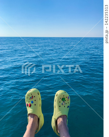 Point of view shot of a pair of legs wearing lime green clogs resting above the calm blue sea on a sunny day outdoors Point of view shot of a pair of legs wearing lime green clogs resting above the calm blue sea on a sunny day outdoors 142235313