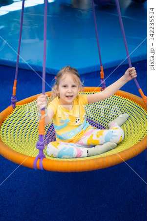 A happy young girl swinging on a colorful circular net chair in a bright indoor play area, laughing with joy during a fun playground adventure in a modern entertainment center A happy young girl swinging on a colorful circular net chair in a bright indoor play area, laughing with joy during a fun playground adventure in a modern entertainment center 142235514