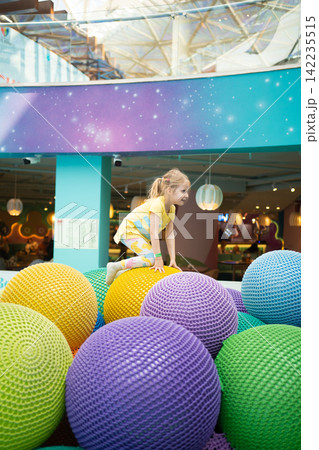 Little blonde girl in a yellow shirt playing on large colorful soft spheres in an indoor modern play area, child having fun and active leisure time in a bright entertainment center for kids Little blonde girl in a yellow shirt playing on large colorful soft spheres in an indoor modern play area, child having fun and active leisure time in a bright entertainment center for kids 142235515