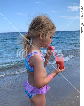 A little blonde girl wearing a colorful swimsuit stands on the sandy beach while drinking a cold red smoothie from a plastic cup with a pink straw against the backdrop of the blue sea A little blonde girl wearing a colorful swimsuit stands on the sandy beach while drinking a cold red smoothie from a plastic cup with a pink straw against the backdrop of the blue sea 142235640