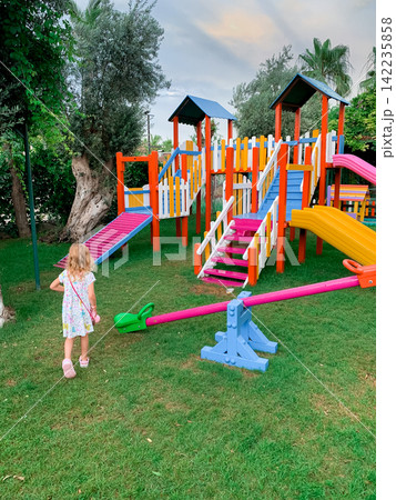 Young girl in a dress walking towards a colorful playground with slides and seesaw in a green garden 142235858