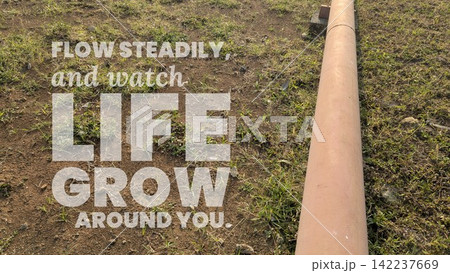 A high angle shot captures inspirational motivational quote text stating "FLOW STEADILY, watch LIFE GROW AROUND YOU." overlaid on a long pipe running across dry grassy ground, soil mixed with small A high angle shot captures inspirational motivational quote text stating "FLOW STEADILY, watch LIFE GROW AROUND YOU." overlaid on a long pipe running across dry grassy ground, soil mixed with small 142237669