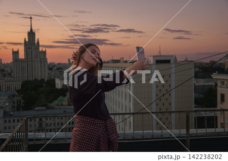 Woman photographs herself with smartphone on Moscow rooftop against warm sunset skyline. Mobile photography, travel lifestyle, urban tourism, self portrait, city exploration, rooftop leisure. Woman photographs herself with smartphone on Moscow rooftop against warm sunset skyline. Mobile photography, travel lifestyle, urban tourism, self portrait, city exploration, rooftop leisure. 142238202
