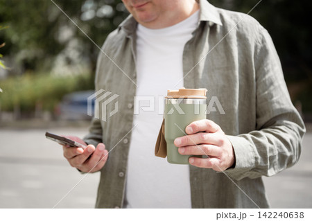 Man holding reusable travel mug and smartphone outdoors 142240638