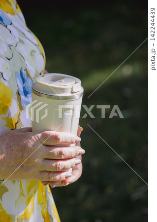 Senior woman hands holding white travel thermo mug in summer garden 142244439