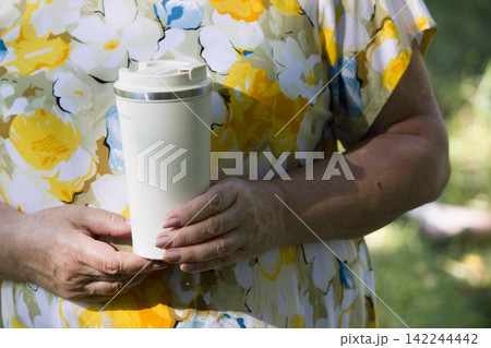 Senior woman holding reusable white travel coffee mug outdoors in park 142244442