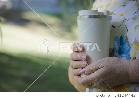 Senior woman holding reusable white travel coffee mug outdoors in sunny park Senior woman holding reusable white travel coffee mug outdoors in sunny park 142244443