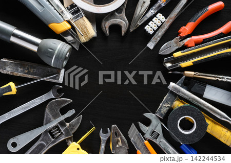 Assorted hand tools and repair supplies arranged around empty center on dark workshop surface Assorted hand tools and repair supplies arranged around empty center on dark workshop surface 142244534