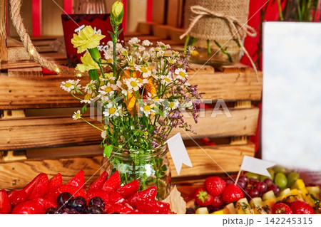 Rustic wooden crate display with a glass jar of fresh wildflowers and chamomile alongside ripe red strawberries 142245315
