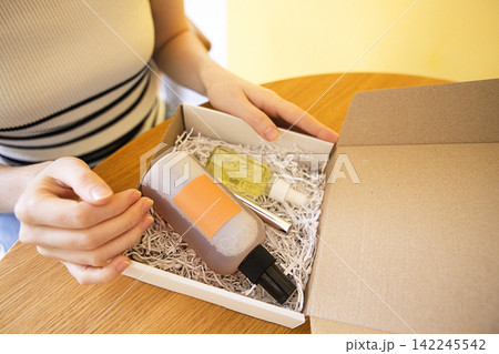 Female hands holding a gift box containing cosmetic bottles with labels, surrounded by shredded paper on a wooden table against a yellow wall Female hands holding a gift box containing cosmetic bottles with labels, surrounded by shredded paper on a wooden table against a yellow wall 142245542