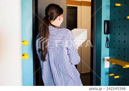 Woman in striped shirt holding a white package while standing at the entrance of a modern apartment with colorful walls and a doorbell visible Woman in striped shirt holding a white package while standing at the entrance of a modern apartment with colorful walls and a doorbell visible 142245566