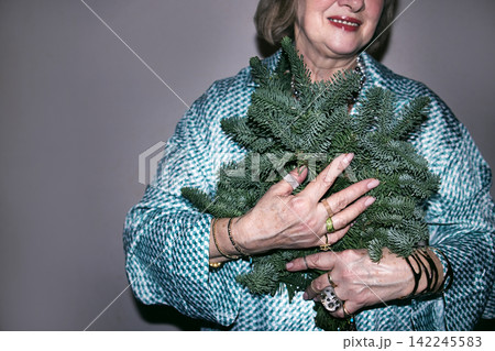 Elderly woman with gray hair wearing a blue patterned shawl holds a bundle of evergreen branches against a neutral background, showcasing festive spirit and seasonal decor Elderly woman with gray hair wearing a blue patterned shawl holds a bundle of evergreen branches against a neutral background, showcasing festive spirit and seasonal decor 142245583
