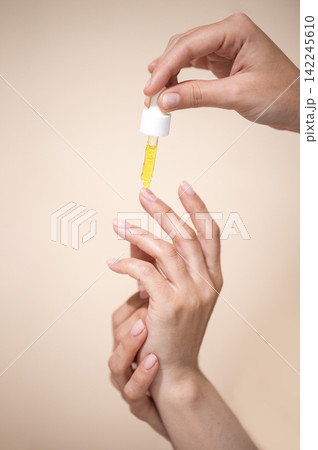 Hands applying yellow serum from dropper onto fingers against a soft beige background, showcasing skincare routine and beauty treatment 142245610