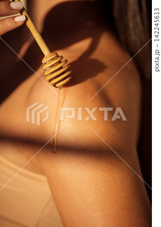 Close-up of honey dripping from wooden dipper onto shoulder of a woman with smooth skin illuminated by soft natural light in a serene indoor setting Close-up of honey dripping from wooden dipper onto shoulder of a woman with smooth skin illuminated by soft natural light in a serene indoor setting 142245613
