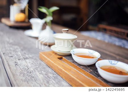 Traditional tea setup with two porcelain cups filled with tea on a wooden tray, surrounded by teapot, bamboo plant, and decorative items in a serene setting Traditional tea setup with two porcelain cups filled with tea on a wooden tray, surrounded by teapot, bamboo plant, and decorative items in a serene setting 142245695