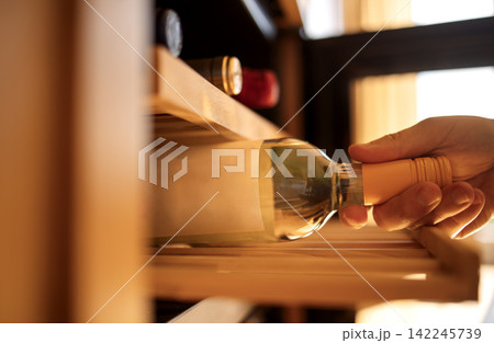 Hand reaching for a bottle of white wine from a wooden wine rack with various bottles visible in the background and natural light illuminating the scene Hand reaching for a bottle of white wine from a wooden wine rack with various bottles visible in the background and natural light illuminating the scene 142245739