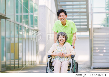 車いすを押す若い男性介護士と高齢者の女性 車いすを押す若い男性介護士と高齢者の女性 142246991