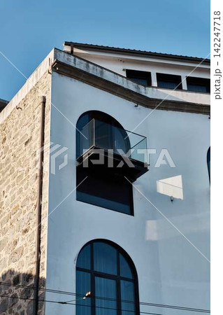 Contemporary building facade with glass balcony, arched windows, stone wall, stucco surfaces, and clean geometric lines beneath bright blue sky with warm sunlight Contemporary building facade with glass balcony, arched windows, stone wall, stucco surfaces, and clean geometric lines beneath bright blue sky with warm sunlight 142247718