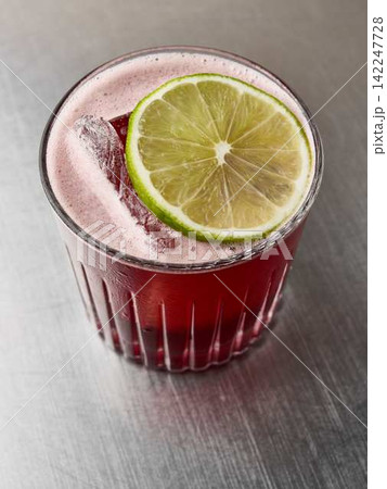 Close up red beverage in glass with lime slice garnish over ice on stainless steel surface high angle, with beverage, drink, red, lime detail, showing slice, garnish, ice, cube context Close up red beverage in glass with lime slice garnish over ice on stainless steel surface high angle, with beverage, drink, red, lime detail, showing slice, garnish, ice, cube context 142247728