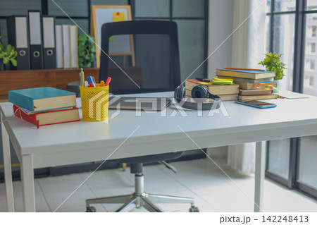 Study desk with a stack of books, black over-ear headphones, a smartphone, sticky notes, laptop, and a small potted plant near a bright window. 142248413