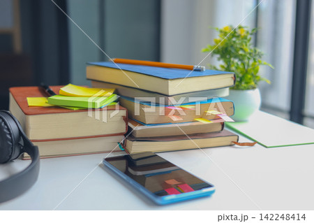 Study desk with a stack of books, black over-ear headphones, a smartphone, sticky notes, and a small potted plant near a bright window. 142248414
