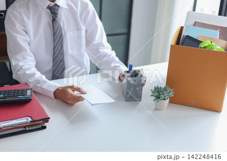 Office worker handing over an envelope, next to a packing box filled with personal belongings. Concept for resignation or leaving a job. Office worker handing over an envelope, next to a packing box filled with personal belongings. Concept for resignation or leaving a job. 142248416