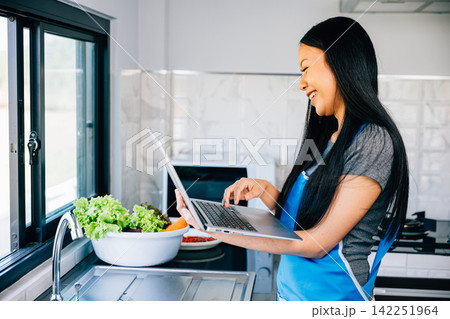 A woman stands in the kitchen happily cooking while using a laptop for video tutorials 142251964
