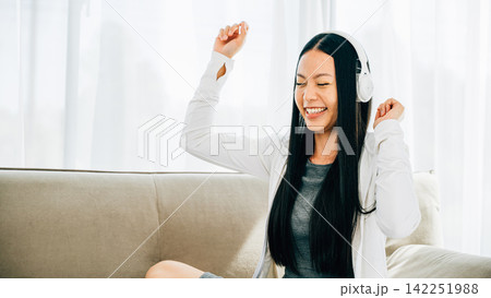Happy woman in headphones holds phone dances and gestures on sofa 142251988