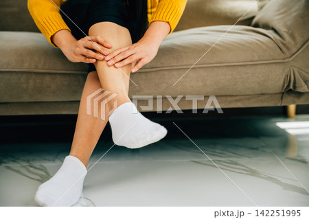Woman sitting on sofa holds her ankle injury expressing pain Woman sitting on sofa holds her ankle injury expressing pain 142251995