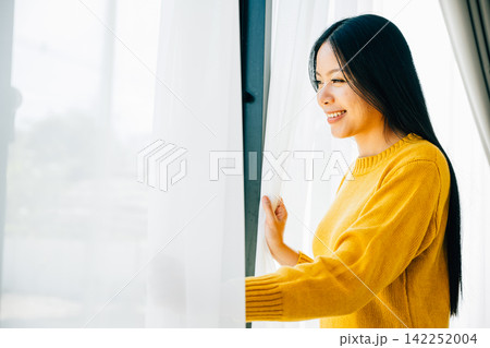 A young woman opens curtains in the early morning smiles at the view feeling relaxed A young woman opens curtains in the early morning smiles at the view feeling relaxed 142252004