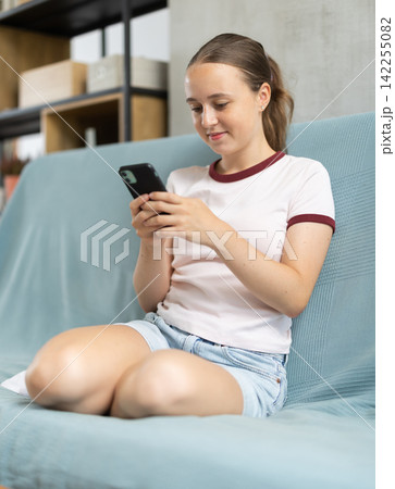 Relaxed teenage girl looking at smartphone while sitting on sofa Relaxed teenage girl looking at smartphone while sitting on sofa 142255082
