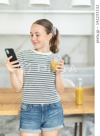 Teenage girl standing in the kitchen, drinking juice and scrolling through social networks on mobile Teenage girl standing in the kitchen, drinking juice and scrolling through social networks on mobile 142255132
