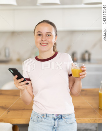 Teenage girl standing in the kitchen, drinking juice and scrolling through social networks on mobile Teenage girl standing in the kitchen, drinking juice and scrolling through social networks on mobile 142255146