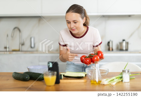 Teenage girl prepares salad in kitchen and looks at smartphone screen Teenage girl prepares salad in kitchen and looks at smartphone screen 142255190