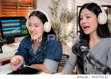 Two young asian woman with headphones speaking on microphone in home studio. 142255213