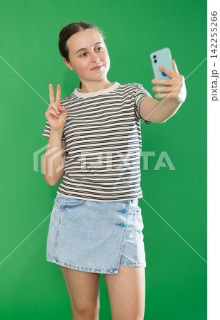 Happy teenage girl in denim skirt takes selfie on a smartphone on green background. Chroma key compositing Happy teenage girl in denim skirt takes selfie on a smartphone on green background. Chroma key compositing 142255266